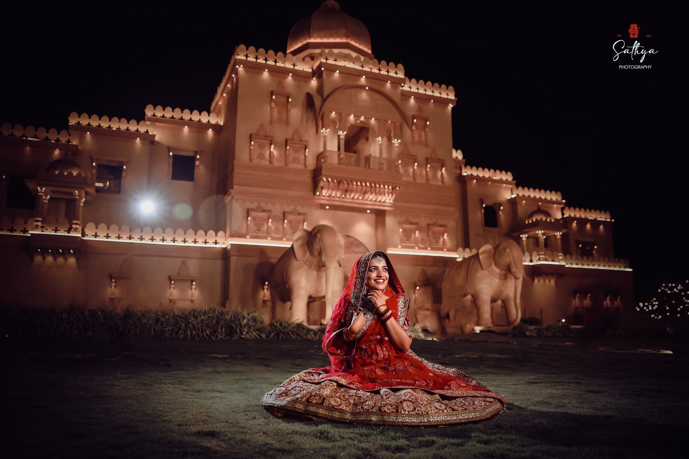 Bride in a red lehenga seated before an illuminated palace