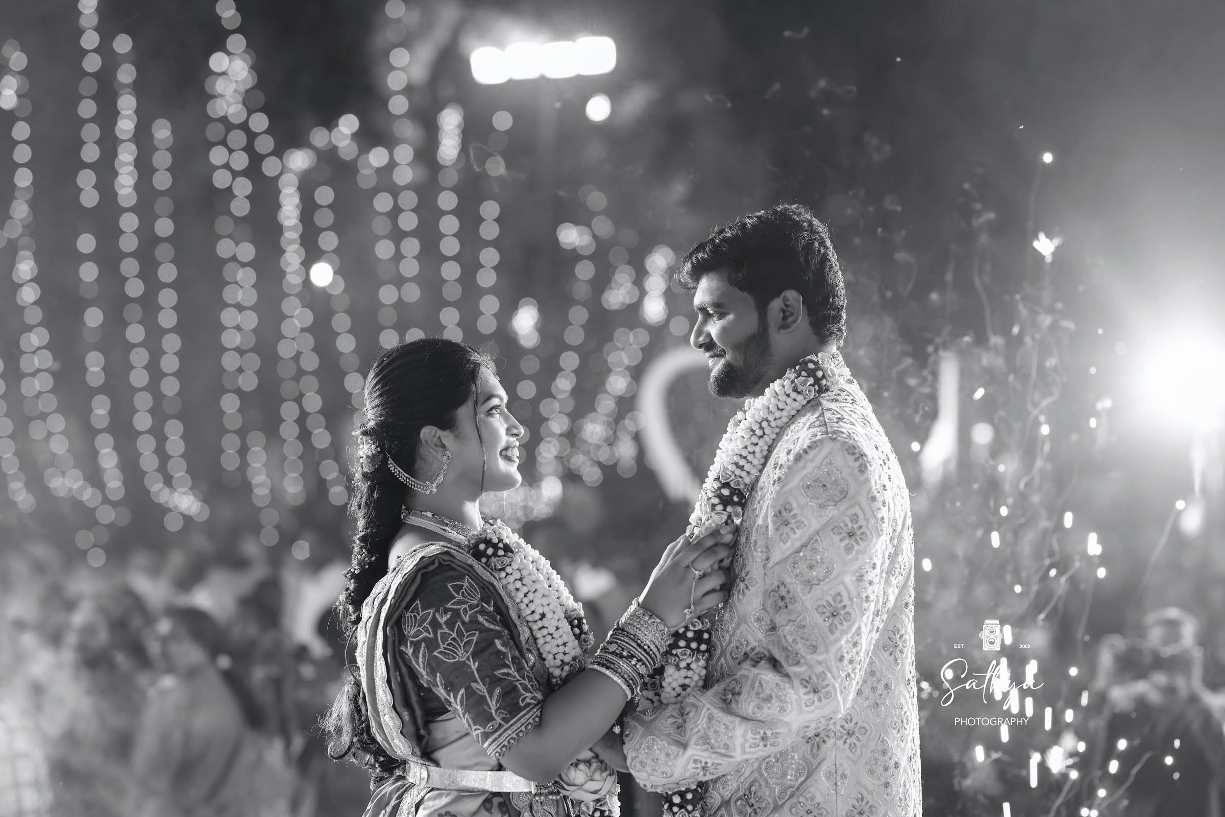 Black-and-white moment of a couple exchanging garlands under lights