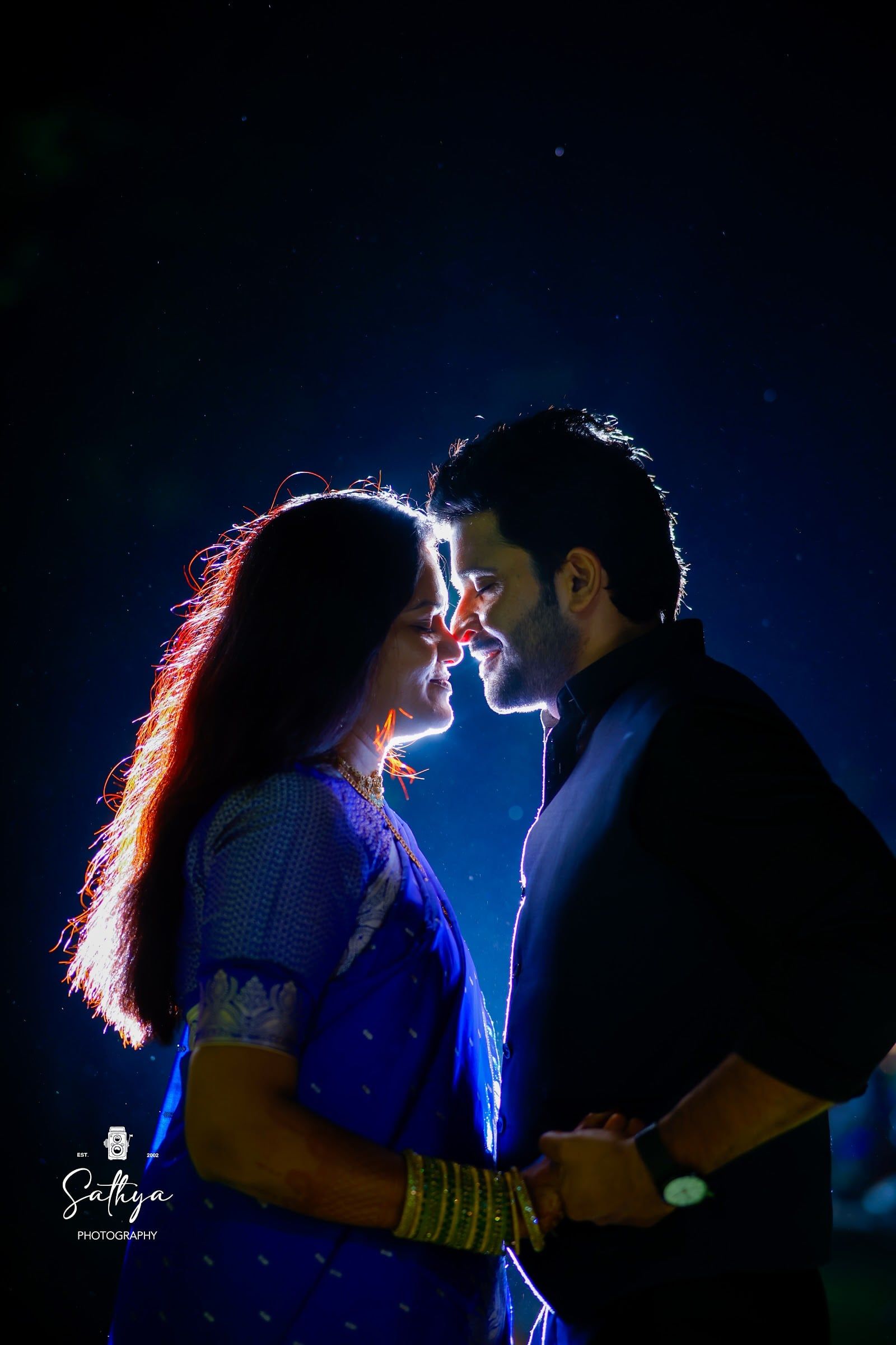 Rim-lit couple forehead to forehead against a deep blue night