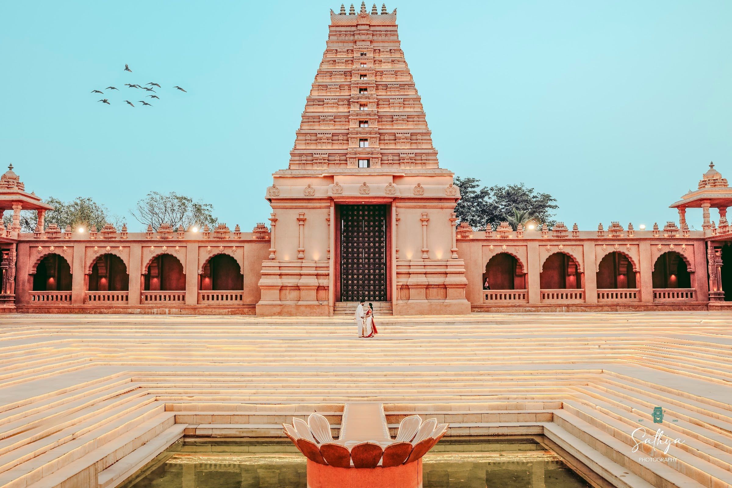 Couple dwarfed by a temple tank and gopuram under a blue sky