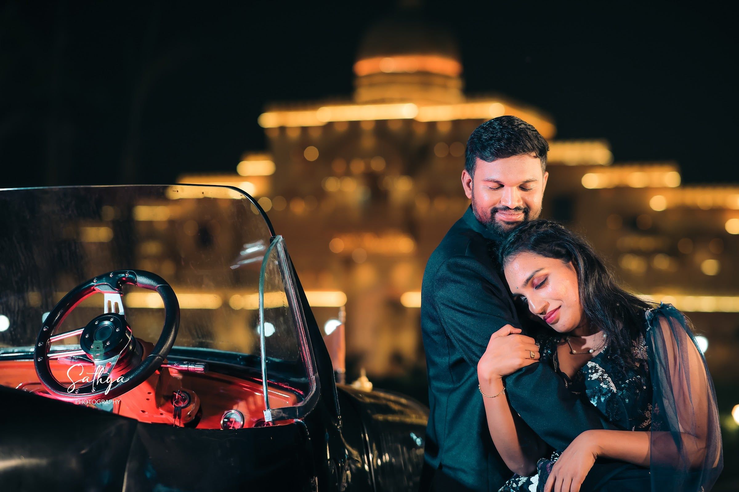 Couple embracing beside a vintage car before a lit palace at night